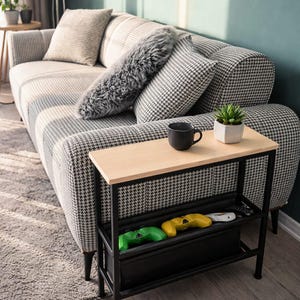 Op de afbeelding: Een woonkamer met een patroonbank, een kleine bijzettafel en een vloerkleed. De bijzettafel heeft een licht houten blad en een zwart metalen frame. Drie gamecontrollers staan in de onderste plank. Een zwarte mok en een plant in een pot staan op de tafel.