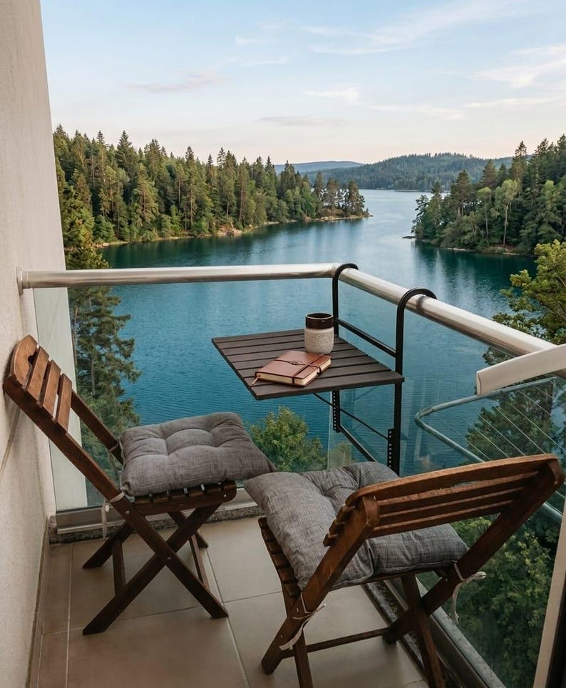 Peut inclure: Sc&egrave;ne de balcon avec vue sur un lac et une for&ecirc;t. Deux chaises pliantes en bois avec des coussins gris font face au lac. Une petite table contient une tasse et un carnet. L'eau est bleu profond.