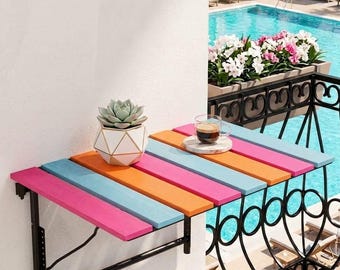 Peut inclure: Une table de balcon murale composée de lattes de bois horizontales peintes en rose vif, bleu clair et orange. La table est fixée par une structure en métal noir et supporte un pot de succulente géométrique blanc et une tasse de café.