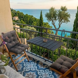Peut inclure: Sc&egrave;ne de balcon avec vue sur la mer. Deux chaises pliantes en bois avec coussins gris et une petite table noire sont sur un sol carrel&eacute;. Le balcon a une balustrade en fer forg&eacute;, et l'arri&egrave;re-plan montre des arbres et l'oc&eacute;an.