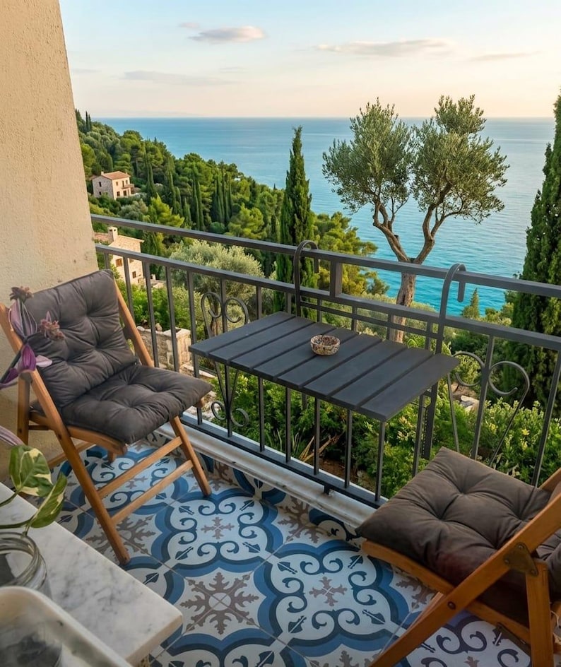 Peut inclure: Sc&egrave;ne de balcon avec vue sur la mer. Deux chaises pliantes en bois avec coussins gris et une petite table noire sont sur un sol carrel&eacute;. Le balcon a une balustrade en fer forg&eacute;, et l'arri&egrave;re-plan montre des arbres et l'oc&eacute;an.