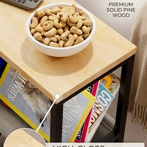Op de afbeelding: Een licht houten bijzettafel met een zwart metalen frame. Een witte kom gevuld met cashewnoten staat bovenop, naast een afstandsbediening. Tijdschriften worden op de plank eronder bewaard. De tekst luidt "HIGH-GLOSS VARNISHED SURFACE".