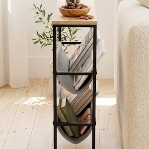 Op de afbeelding: Een smalle bijzettafel met een licht houten blad en een zwart metalen frame. Het heeft een ingebouwd tijdschriftenrek met grijze stoffen banden en verstelbare poten voor stabiliteit. Een cafeti&egrave;re, houten kom en boeken zijn te zien.