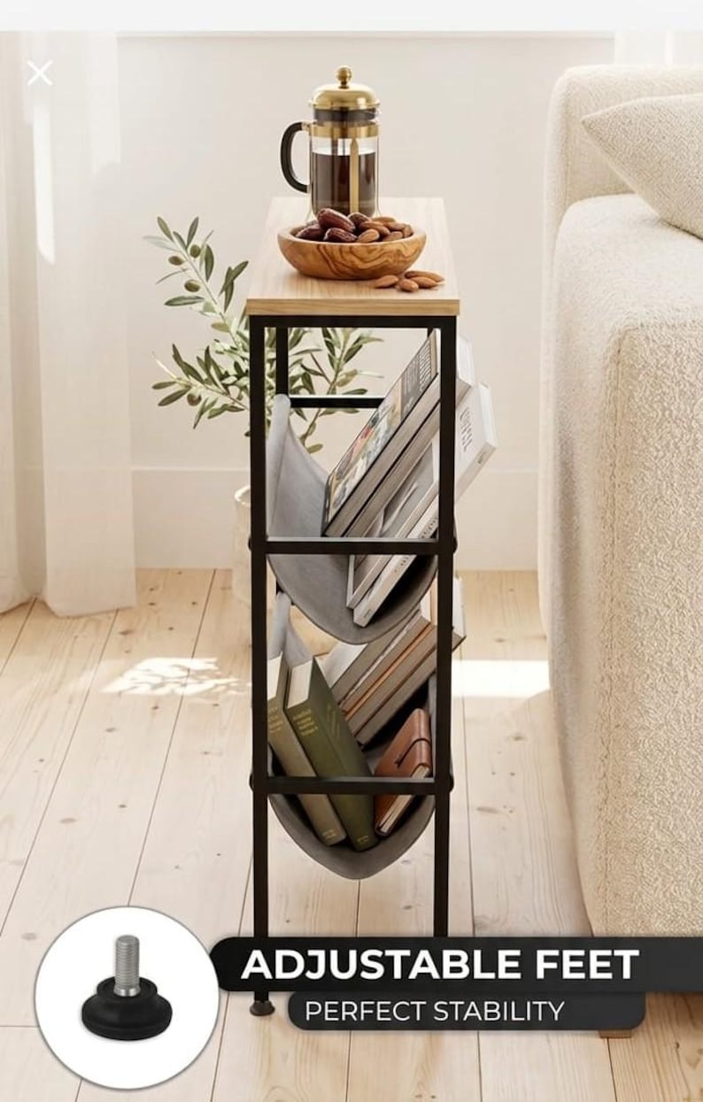 Op de afbeelding: Een smalle bijzettafel met een licht houten blad en een zwart metalen frame. Het heeft een ingebouwd tijdschriftenrek met grijze stoffen banden en verstelbare poten voor stabiliteit. Een cafeti&egrave;re, houten kom en boeken zijn te zien.
