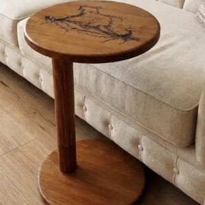 Pode incluir: Uma mesa lateral redonda de madeira com uma &uacute;nica coluna de suporte cil&iacute;ndrica e uma base circular. O tampo apresenta um acabamento de gr&atilde;o de madeira natural com uma incrusta&ccedil;&atilde;o de resina escura ramificada que cria um padr&atilde;o org&acirc;nico.
