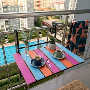May include: A multi-coloured folding balcony table with pink, blue, and orange planks. Two cups of coffee sit on the table, alongside a small plant and a vase of greenery. The table is attached to a metal railing. The table is a great space saver.