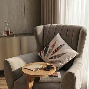 Puede incluir: Escena de salón acogedora con un sillón gris y un cojín decorativo con diseños de plumas. Una pequeña mesa auxiliar de madera sostiene una taza de café y libros, incluyendo "Kinfolk Minimalist Homes".