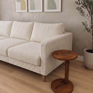 Pode incluir: Um sof&aacute; de cor creme estofado em tecido texturizado e irregular, com uma estrutura retangular e pernas de madeira clara, posicionado ao lado de uma pequena mesa lateral redonda de madeira castanho-escura com base central e veios vis&iacute;veis.