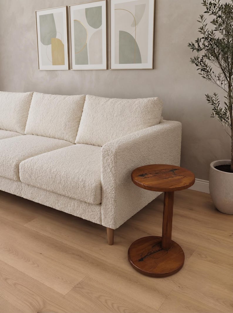Pode incluir: Um sof&aacute; de cor creme estofado em tecido texturizado e irregular, com uma estrutura retangular e pernas de madeira clara, posicionado ao lado de uma pequena mesa lateral redonda de madeira castanho-escura com base central e veios vis&iacute;veis.