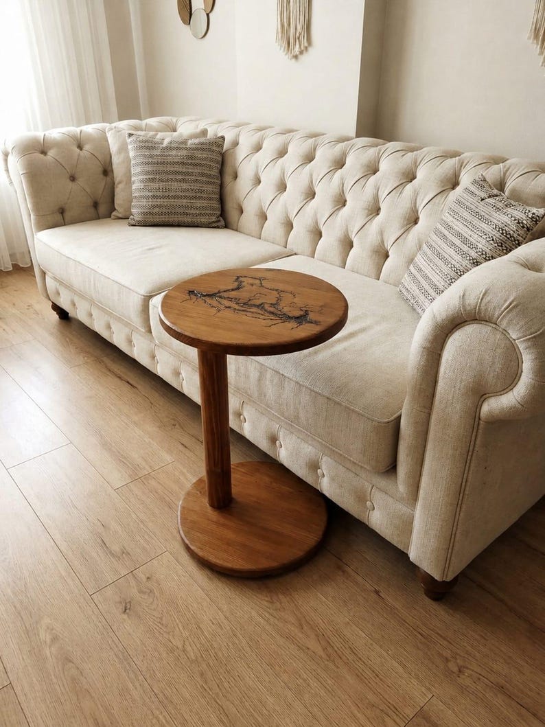 Pode incluir: Uma mesa lateral redonda de madeira com uma base de pedestal central &uacute;nica e um tampo circular com padr&otilde;es fractais escuros e ramificados. A mesa est&aacute; posicionada em frente a um sof&aacute; bege capiton&ecirc; com duas almofadas estampadas.