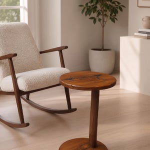 Pode incluir: Uma mesa lateral redonda de madeira com acabamento de gr&atilde;o natural e incrusta&ccedil;&otilde;es de resina escura de formas org&acirc;nicas no topo e na base. A mesa possui uma coluna central cil&iacute;ndrica que liga o tampo circular &agrave; base circular.