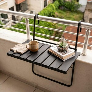 Peut inclure: Table de balcon en bois noir avec cadre en m&eacute;tal, supportant une tasse beige, un tissu pli&eacute;, un petit terrarium et des livres. La table est fix&eacute;e &agrave; une balustrade en m&eacute;tal, avec vue sur la verdure.