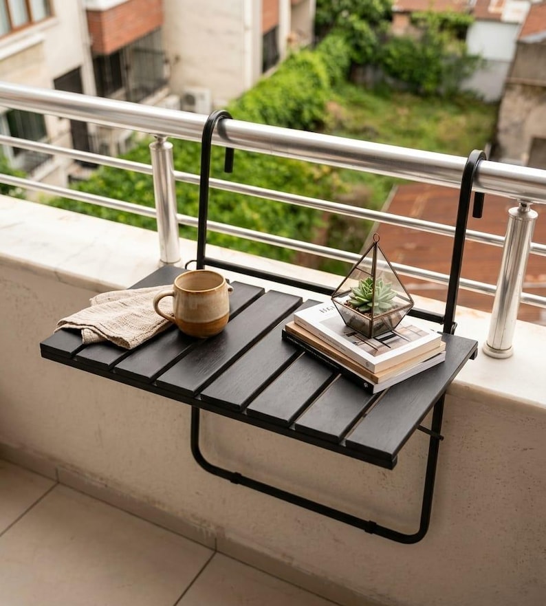 Peut inclure: Table de balcon en bois noir avec cadre en m&eacute;tal, supportant une tasse beige, un tissu pli&eacute;, un petit terrarium et des livres. La table est fix&eacute;e &agrave; une balustrade en m&eacute;tal, avec vue sur la verdure.