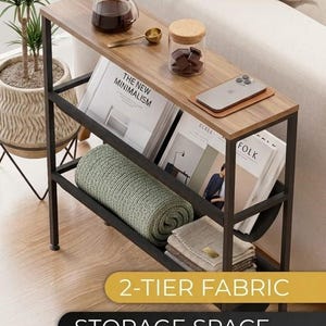 Op de afbeelding: Een bijzettafel met twee niveaus, met een houten blad en een zwart metalen frame. De bovenste plank bevat een koffiezetapparaat, een klein potje en een telefoon. De onderste plank bevat opgerolde dekens en boeken. De tekst "2-TIER FABRIC STORAGE SPACE" wordt weergegeven.