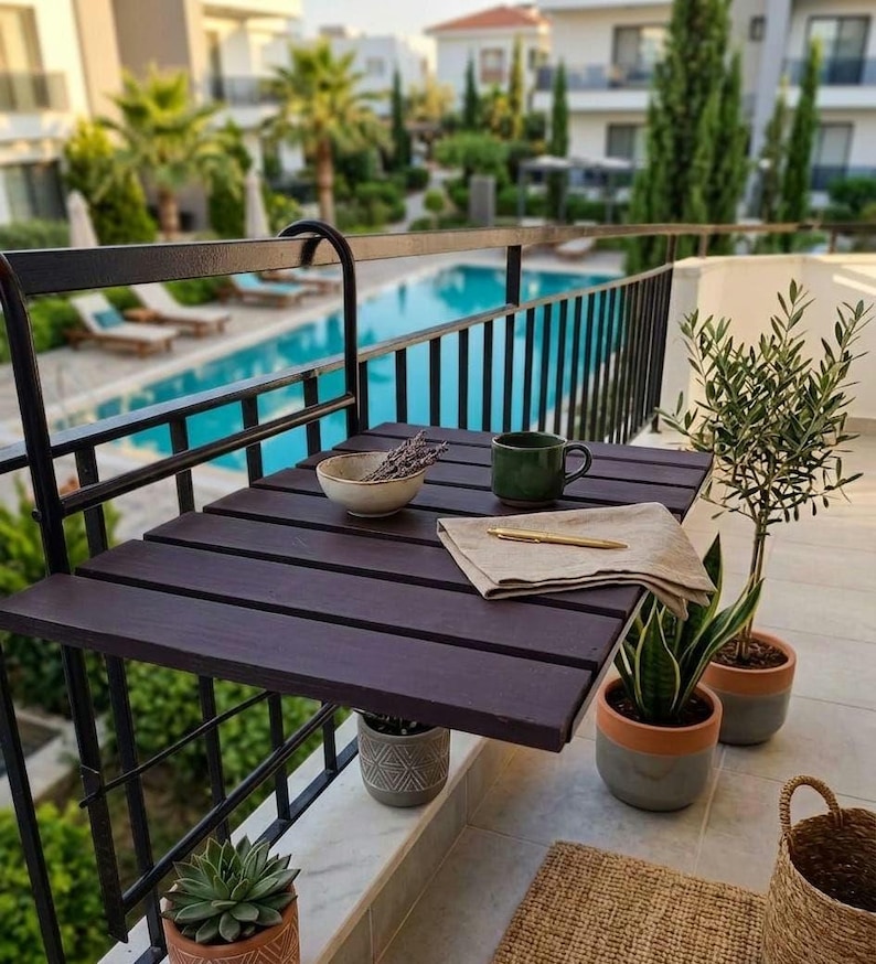 Peut inclure: Une table de balcon pliable, marron fonc&eacute;, avec un cadre en m&eacute;tal noir. Sur la table, un petit bol de lavande, une tasse verte, une serviette beige et un stylo. Des plantes en pot et un panier en osier sont &eacute;galement visibles, avec une piscine en arri&egrave;re-plan.