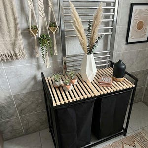 May include: A black metal laundry sorter with two fabric bags and a slatted wooden top. On the top surface sits a white geometric vase with dried pampas grass, three small air plants in concrete planters, and a black diffuser with cork base.