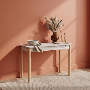 May include: A rectangular console table with light wood legs and a white marble-patterned apron. On the surface sits a dark brown glass vase filled with dried pampas grass, a white ceramic bowl, a folded beige cloth, and two gold candle holders.