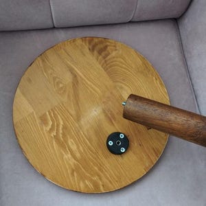 Pode incluir: Um tampo de mesa circular de madeira castanho-clara com um padr&atilde;o de gr&atilde;o vis&iacute;vel. Uma perna de madeira cil&iacute;ndrica com um parafuso de metal no topo est&aacute; posicionada &agrave; direita. Uma pequena placa de montagem preta circular est&aacute; fixada.