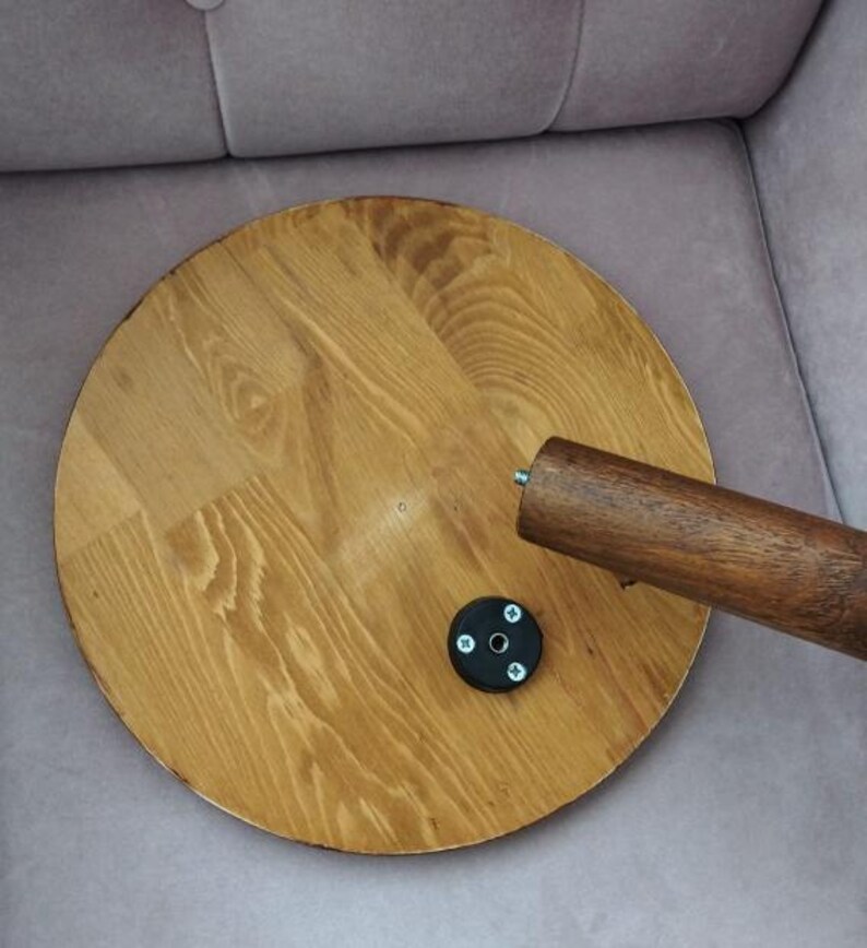 Pode incluir: Um tampo de mesa circular de madeira castanho-clara com um padr&atilde;o de gr&atilde;o vis&iacute;vel. Uma perna de madeira cil&iacute;ndrica com um parafuso de metal no topo est&aacute; posicionada &agrave; direita. Uma pequena placa de montagem preta circular est&aacute; fixada.