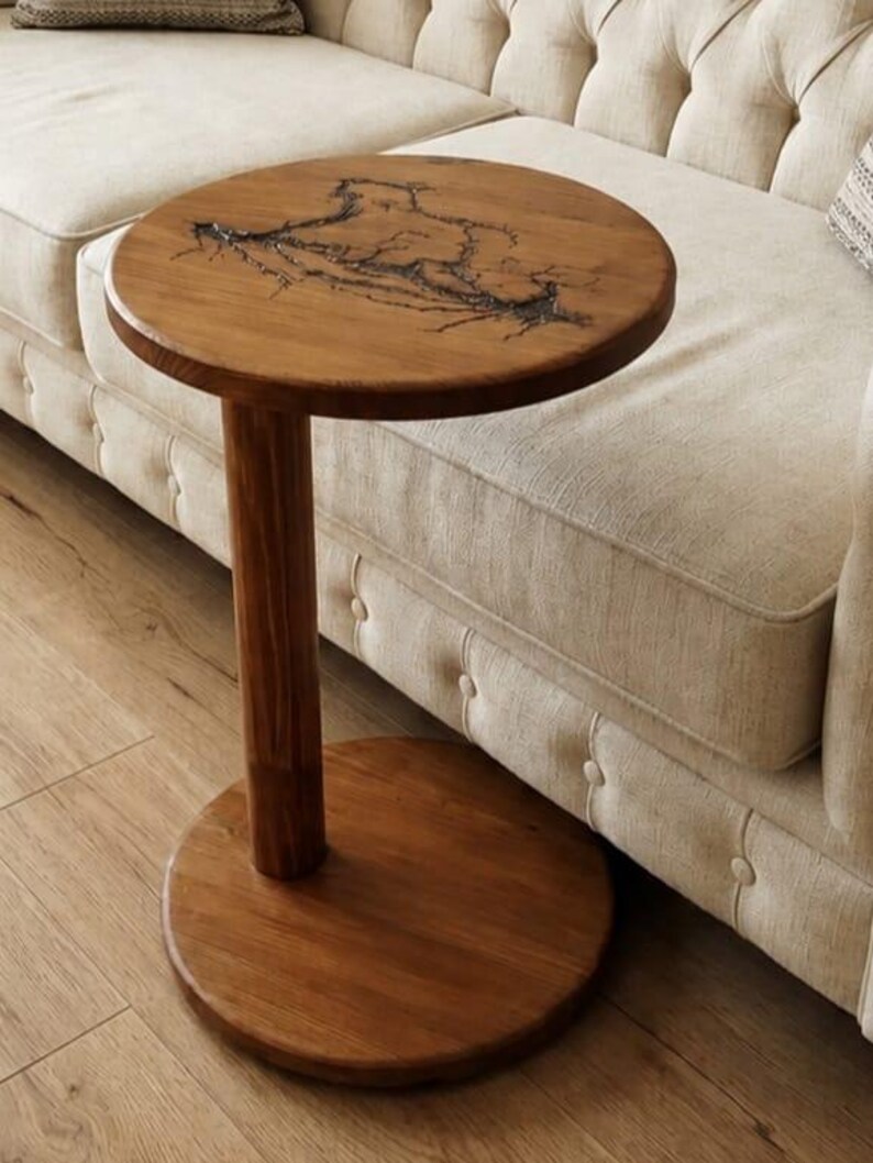 Pode incluir: Uma mesa lateral redonda de madeira com uma base de pedestal cil&iacute;ndrica e uma placa de base circular. O tampo da mesa apresenta um padr&atilde;o de queima fractal escuro e ramificado sobre o veio natural da madeira, com um acabamento liso e polido.