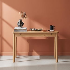 May include: A light-toned wooden desk with four cylindrical legs and a single drawer. On the desk are three stacked books, a dark gray abstract ceramic vase with a dried flower, a shallow dark bowl with stones, a brown notebook, and a black mug.