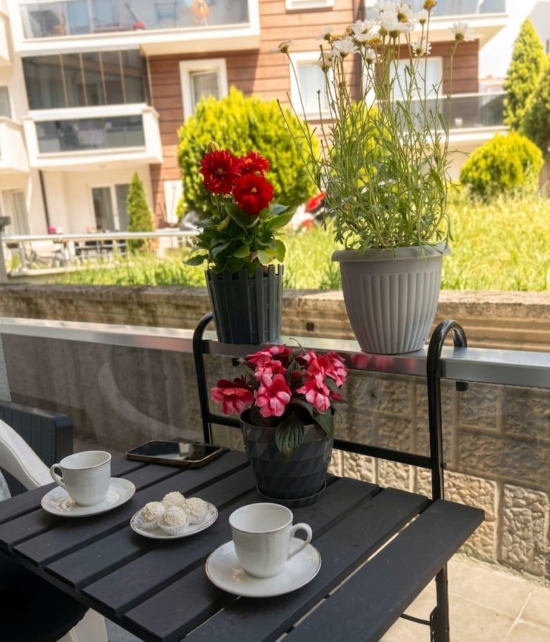 Peut inclure: Une table noire de jardin avec deux tasses &agrave; th&eacute; et soucoupes blanches, une assiette de friandises blanches et un smartphone. La table est orn&eacute;e de fleurs rouges et roses en pot et de marguerites. Une balustrade en m&eacute;tal noir soutient les plantes en pot.