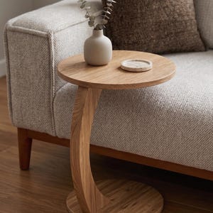 Può includere: Un tavolino rotondo in legno con base di supporto curva. Sul piano del tavolo si trovano un piccolo vaso in ceramica beige opaco contenente un rametto di eucalipto essiccato e un sottobicchiere rotondo in pietra chiara dalla superficie porosa.