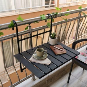 Peut inclure: Une table de balcon noire &agrave; lattes avec une tasse et une soucoupe, une petite plante succulente en pot et un carnet avec un stylo. La table est fix&eacute;e &agrave; une balustrade en m&eacute;tal avec un support noir. De la verdure est drap&eacute;e sur la balustrade.