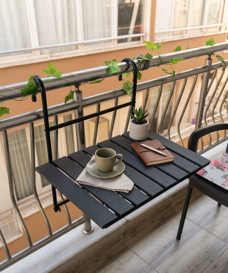 Peut inclure: Une table de balcon noire &agrave; lattes avec une tasse et une soucoupe, une petite plante succulente en pot et un carnet avec un stylo. La table est fix&eacute;e &agrave; une balustrade en m&eacute;tal avec un support noir. De la verdure est drap&eacute;e sur la balustrade.