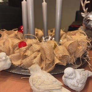 May include: A festive Advent wreath with four gray candles, surrounded by a burlap base and decorative red ornaments. Two small, stone-like bird figurines are in the foreground, creating a seasonal display.