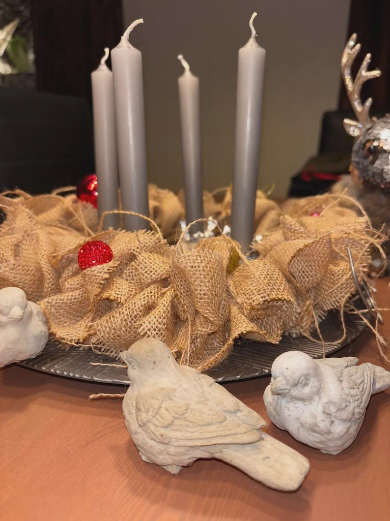 May include: A festive Advent wreath with four gray candles, surrounded by a burlap base and decorative red ornaments. Two small, stone-like bird figurines are in the foreground, creating a seasonal display.