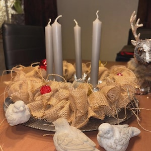 May include: A rustic Advent wreath with four gray candles, surrounded by burlap and red ornaments. Three small, light-colored bird figurines are in the foreground. The wreath sits on a metal tray, creating a cozy, seasonal display.