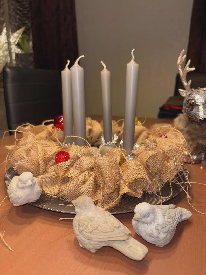 May include: A rustic Advent wreath with four gray candles, surrounded by burlap and red ornaments. Three small, light-colored bird figurines are in the foreground. The wreath sits on a metal tray, creating a cozy, seasonal display.