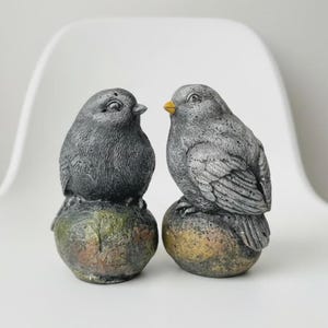 May include: Two gray bird figurines with textured plumage, each perched on a rounded, mottled stone-like base. The bird on the right features a small, distinct yellow beak, while both figures exhibit a weathered, speckled finish throughout.