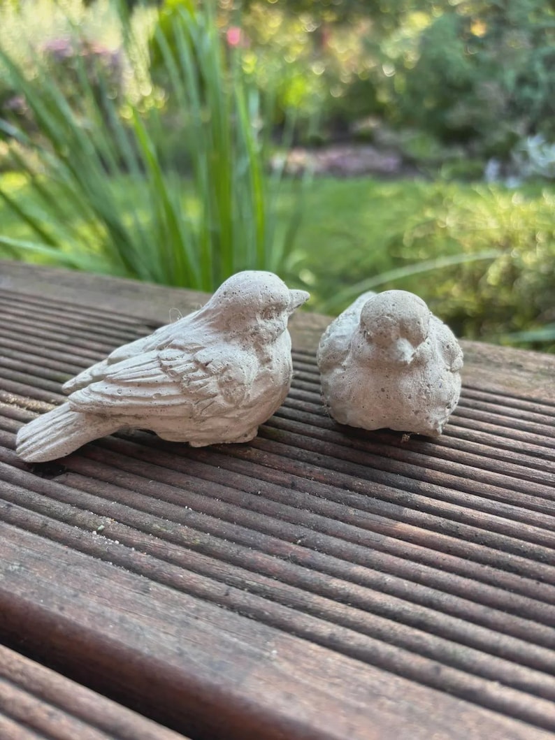 May include: Two small, light gray bird figurines. One bird is standing, the other is sitting. The birds are made of a textured material and are placed on a wooden surface. The background is blurred with green foliage.