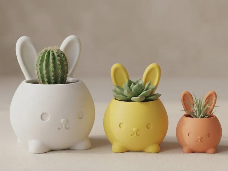 May include: Three round ceramic planters shaped like rabbits in descending sizes. The largest is white with a green cactus, the medium is yellow with a green succulent, and the smallest is orange with a spiky air plant. Each has ears and a face.