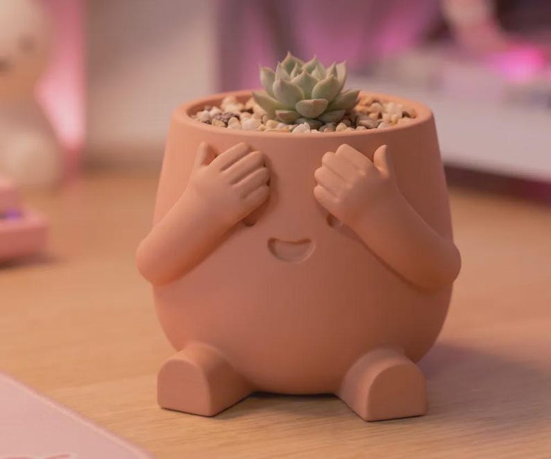 May include: A peach-colored ceramic planter shaped like a figure with two hands covering its eyes and a small smiling mouth. The planter contains a small green succulent plant nestled in light-colored gravel, with two small feet at the base.