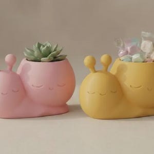 May include: Two snail-shaped planters, one in matte pink and one in matte yellow. Each features a smooth, rounded body with two eye stalks and a carved, smiling face with closed eyes. The pink planter holds a green succulent, while the yellow one holds small candies in clear wrappers.