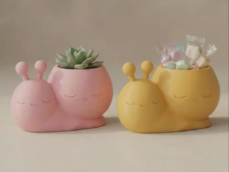 May include: Two snail-shaped planters, one in matte pink and one in matte yellow. Each features a smooth, rounded body with two eye stalks and a carved, smiling face with closed eyes. The pink planter holds a green succulent, while the yellow one holds small candies in clear wrappers.