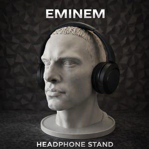 May include: A light grey bust of a person with short hair wearing black over-ear headphones. The word EMINEM is printed in white at the top, and the words HEADPHONE STAND are printed in white at the bottom of the display piece.