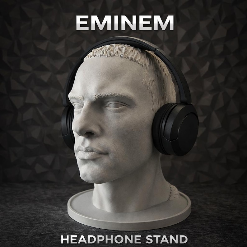 May include: A light grey bust of a person with short hair wearing black over-ear headphones. The word EMINEM is printed in white at the top, and the words HEADPHONE STAND are printed in white at the bottom of the display piece.