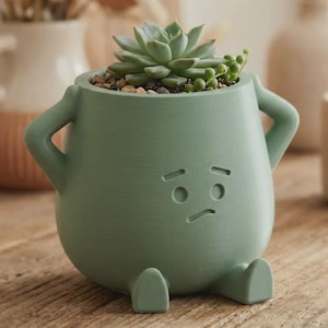 May include: A sage green ceramic planter shaped like a seated figure with arms akimbo and a sad facial expression. The planter contains a small green succulent plant nestled in soil and small pebbles, featuring two small feet at the base.