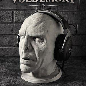 May include: A pale, textured bust of Voldemort featuring prominent facial features and a flat nose, wearing black over-ear headphones with a silver headband. The word VOLDEMORT is printed in white capital letters above the head on a dark background.