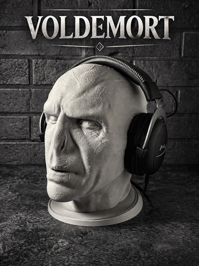 May include: A pale, textured bust of Voldemort featuring prominent facial features and a flat nose, wearing black over-ear headphones with a silver headband. The word VOLDEMORT is printed in white capital letters above the head on a dark background.