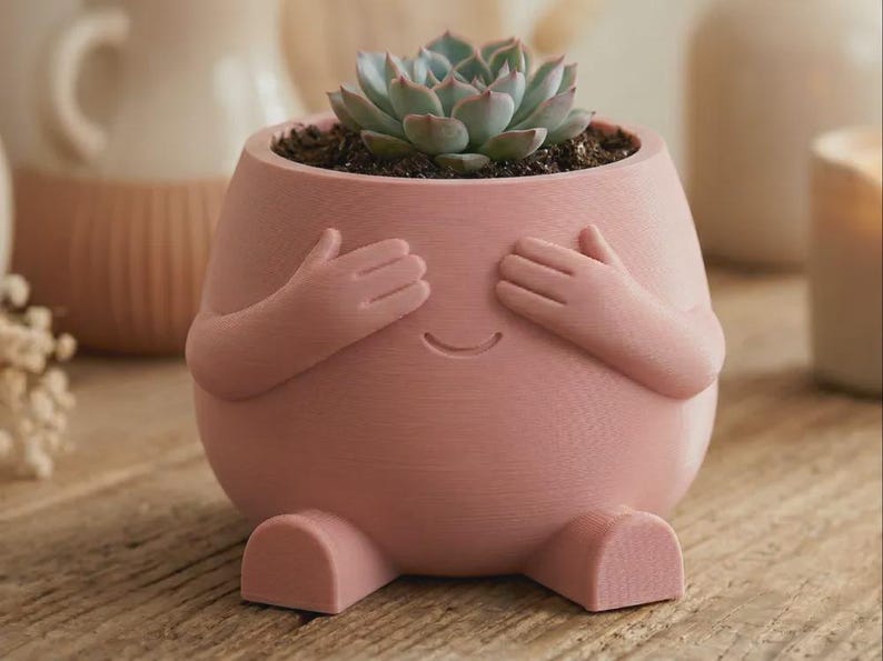 May include: A matte pink planter shaped like a rounded figure with two arms covering its eyes and two small feet at the base. The planter features a simple etched smile and contains a green succulent plant potted in dark soil within the top opening.