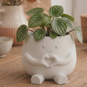 May include: A matte white ceramic planter shaped like a small figure with arms forming a heart shape in front of its chest. The planter features a simple etched face with two circular eyes and a curved mouth, and it contains a watermelon peperomia plant.