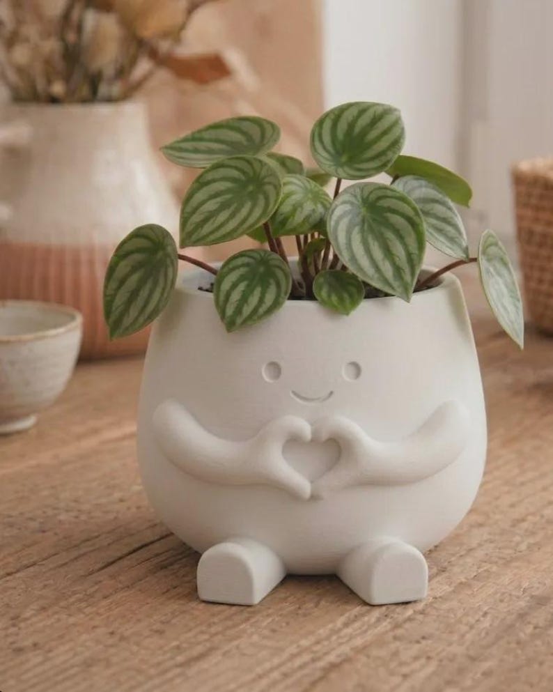 May include: A matte white ceramic planter shaped like a small figure with arms forming a heart shape in front of its chest. The planter features a simple etched face with two circular eyes and a curved mouth, and it contains a watermelon peperomia plant.