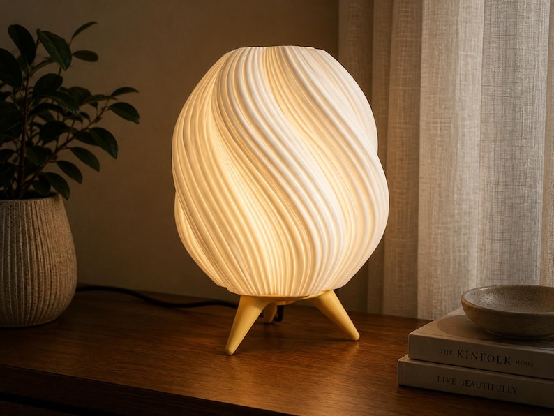 K&ouml;nnte beinhalten: Eine wei&szlig;e Tischlampe mit einem abgerundeten, spiralf&ouml;rmig gerippten Lampenschirm, der ein warmes Licht ausstrahlt. Die Lampe steht auf drei kurzen, hellen, konisch zulaufenden Beinen, die den Hauptk&ouml;rper von der Oberfl&auml;che abheben.