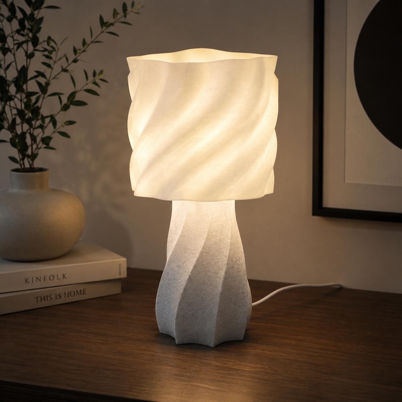 K&ouml;nnte beinhalten: Eine wei&szlig;e Tischleuchte mit einem strukturierten, spiralf&ouml;rmigen Fu&szlig; und einem passenden zylindrischen Lampenschirm mit wellenf&ouml;rmigem, gedrehtem Oberfl&auml;chenmuster. Die Lampe strahlt ein warmes, sanftes Licht aus, das die komplexe Struktur betont.