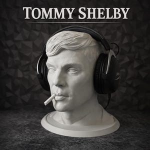 May include: A white bust of a man with a cigarette in his mouth, wearing black over-ear headphones. The text TOMMY SHELBY is printed in white above the head. The sculpture features detailed facial features and hair, resting on a circular base.